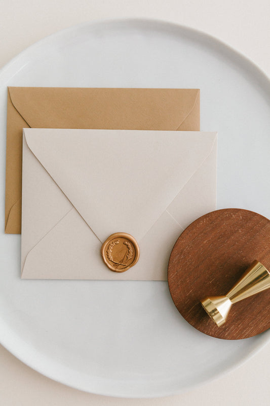 Brass wax seal stamp on a wooden dish beside envelopes with a gold laurel wax seal, styled on a white ceramic plate.