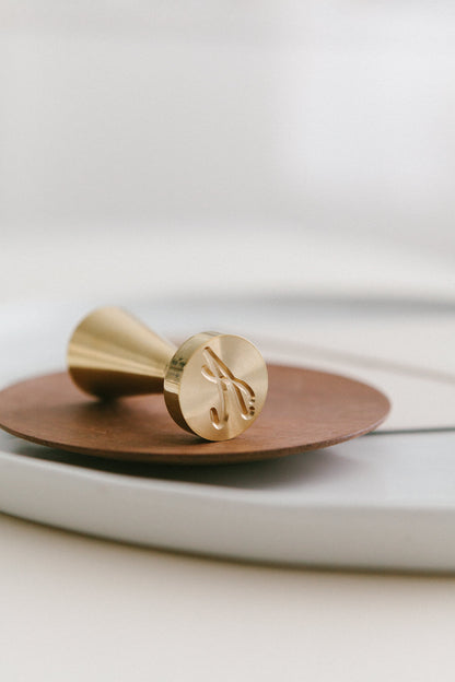 A brass wax seal stamp with A Monogram crest design styled on a wooden and white ceramic plate