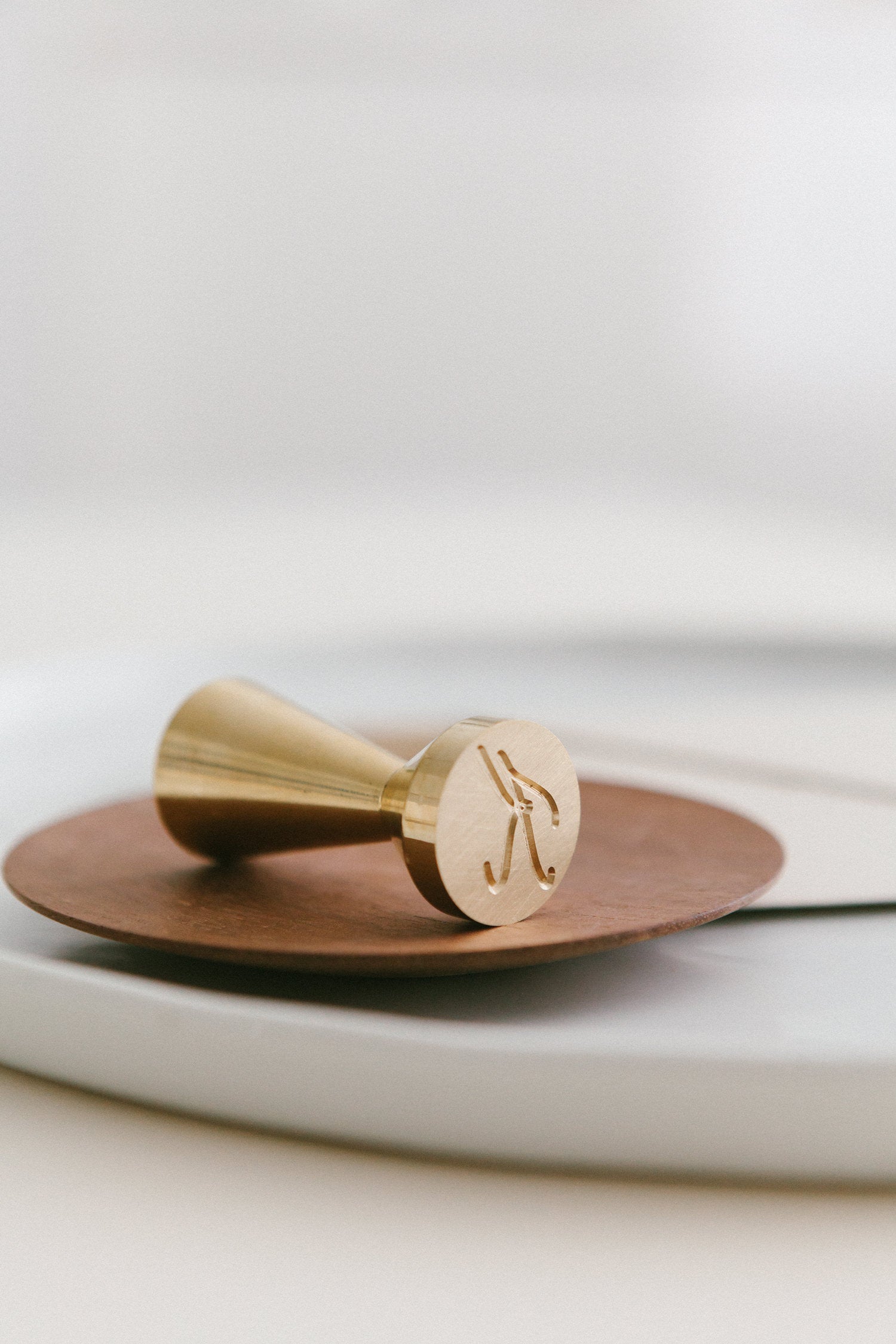 A brass wax seal stamp with a K monogram design styled on a wooden and white ceramic plate