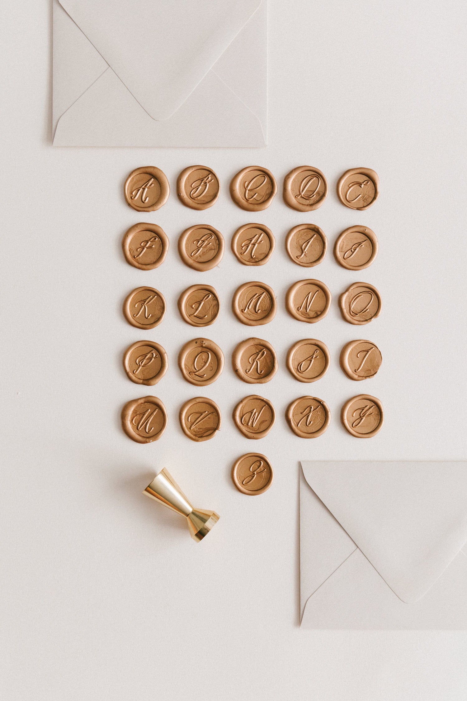 Gold monogram wax seals arranged in alphabetical order with a gold wax seal stamp and two ivory envelopes on a neutral background.