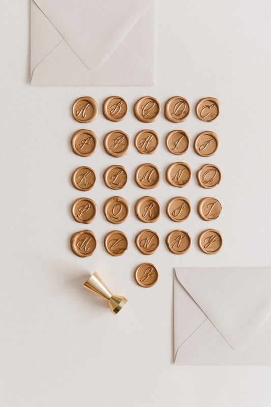 Gold monogram wax seals arranged in alphabetical order with a gold wax seal stamp and two ivory envelopes on a neutral background.