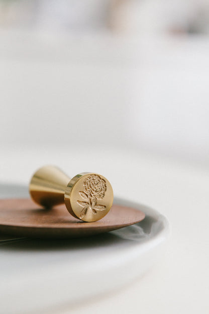 A brass wax seal stamp with heirloom peony design styled on a wooden and white ceramic plate