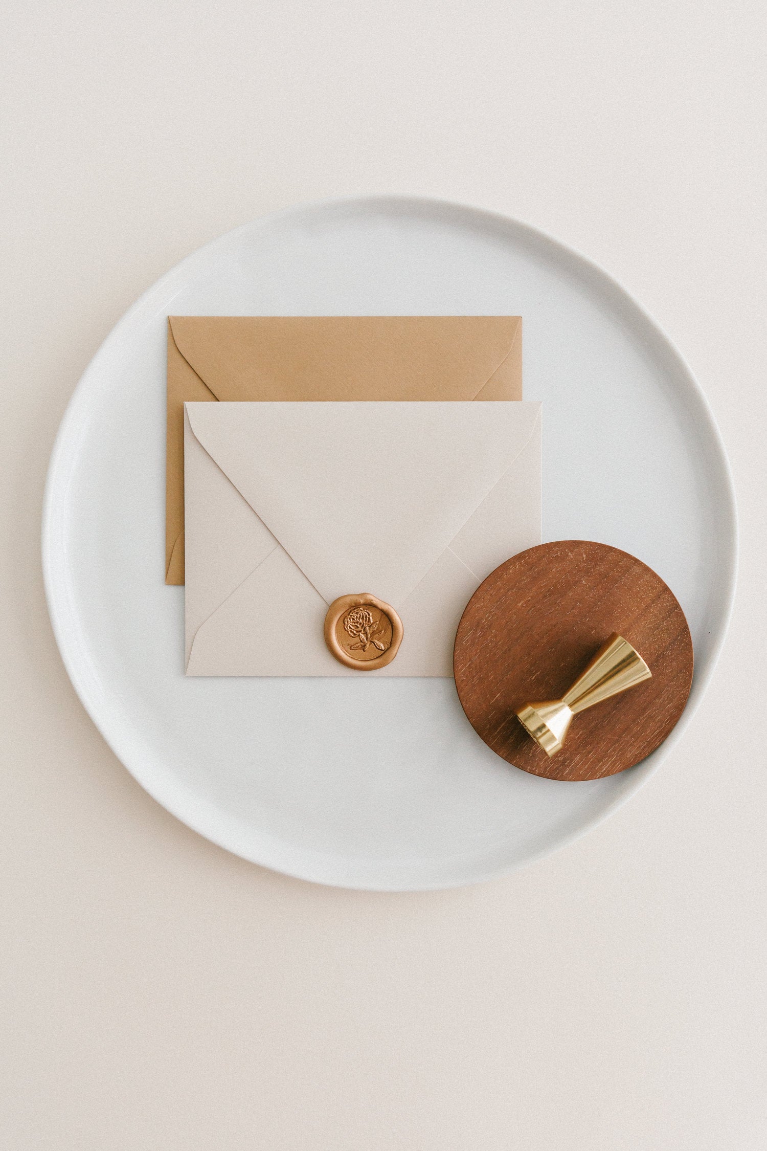 Brass wax seal stamp on a wooden dish beside envelopes with a gold heirloom peony wax seal, styled on a white ceramic plate.