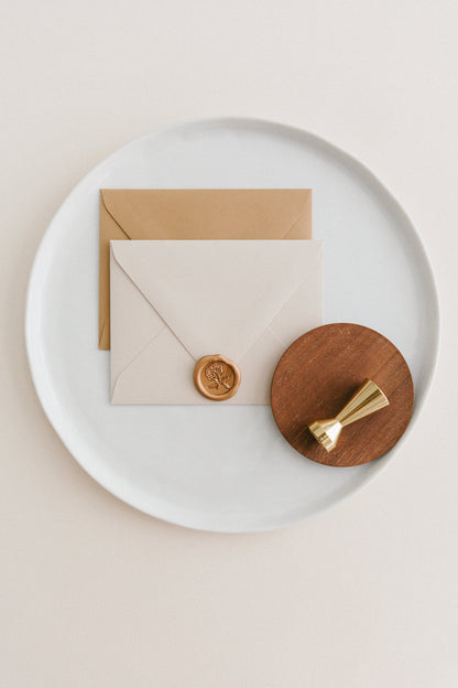Brass wax seal stamp on a wooden dish beside envelopes with a gold heirloom peony wax seal, styled on a white ceramic plate.