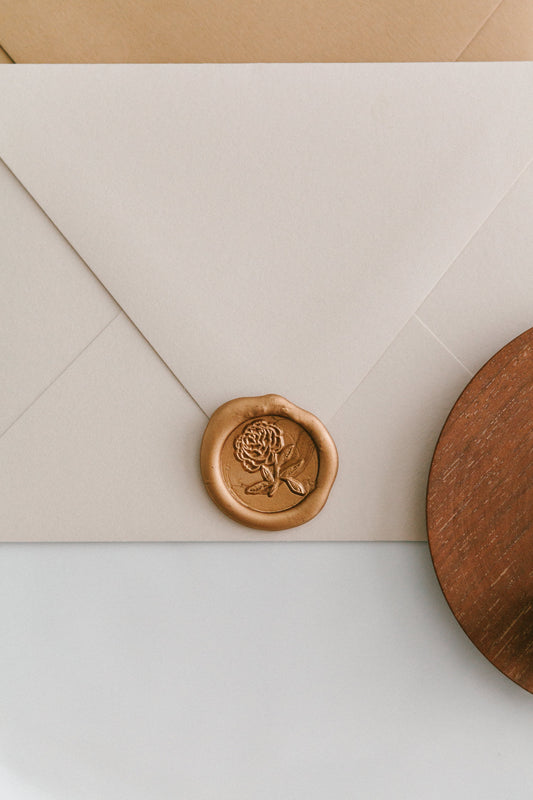A golden heirloom peony wax seal, styled on a beige sealed envelope and a wooden dish on the side