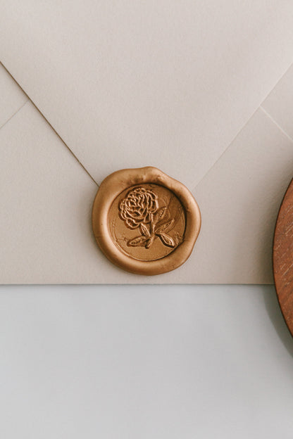 A close-up of a golden heirloom peony wax seal, styled on a beige sealed envelope on a white surface