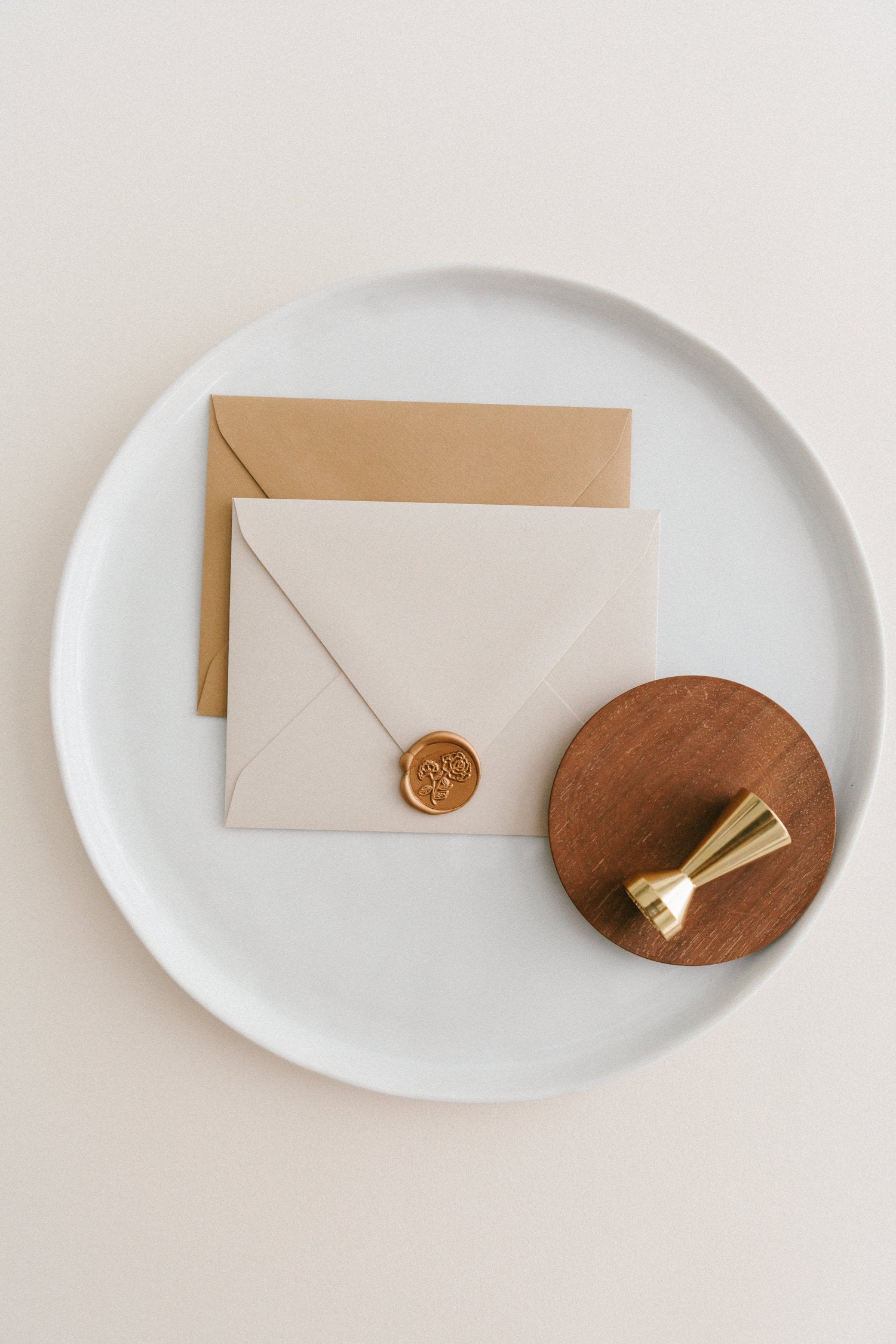 Brass wax seal stamp on a wooden dish beside envelopes with a gold heirloom peony wax seal, styled on a white ceramic plate.