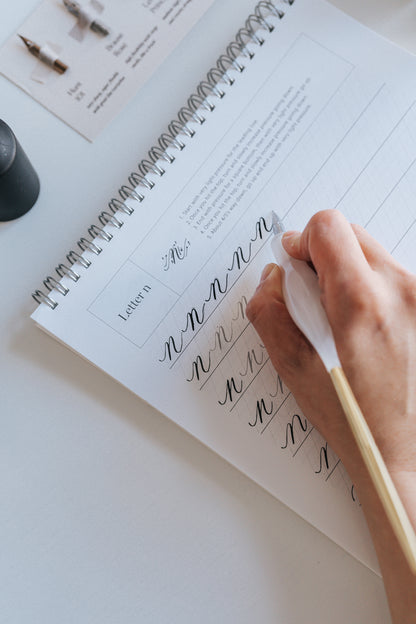 A hand holding a straight pen with a gold tail, practicing modern calligraphy on letter "n" in a calligraphy workbook
