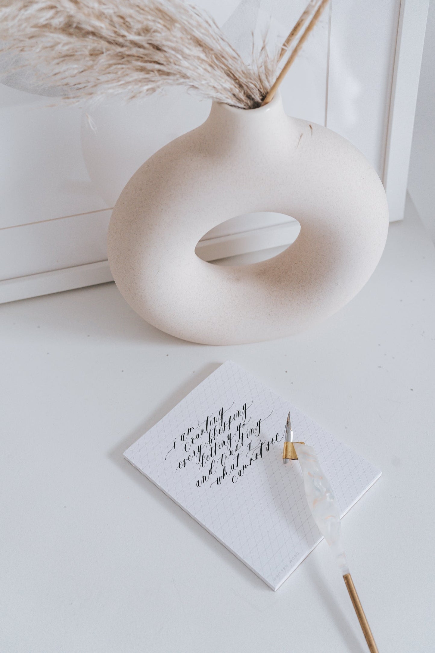 Calligraphy pen on slanted grid notepad with decorative vase and pampas grass in the background