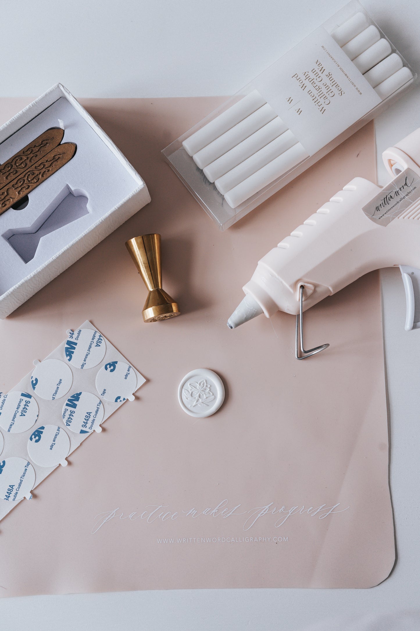 Flat lay of wax seal supplies, including a brass wax seal stamp, white sealing wax sticks, adhesives, and a glue gun on a mat.