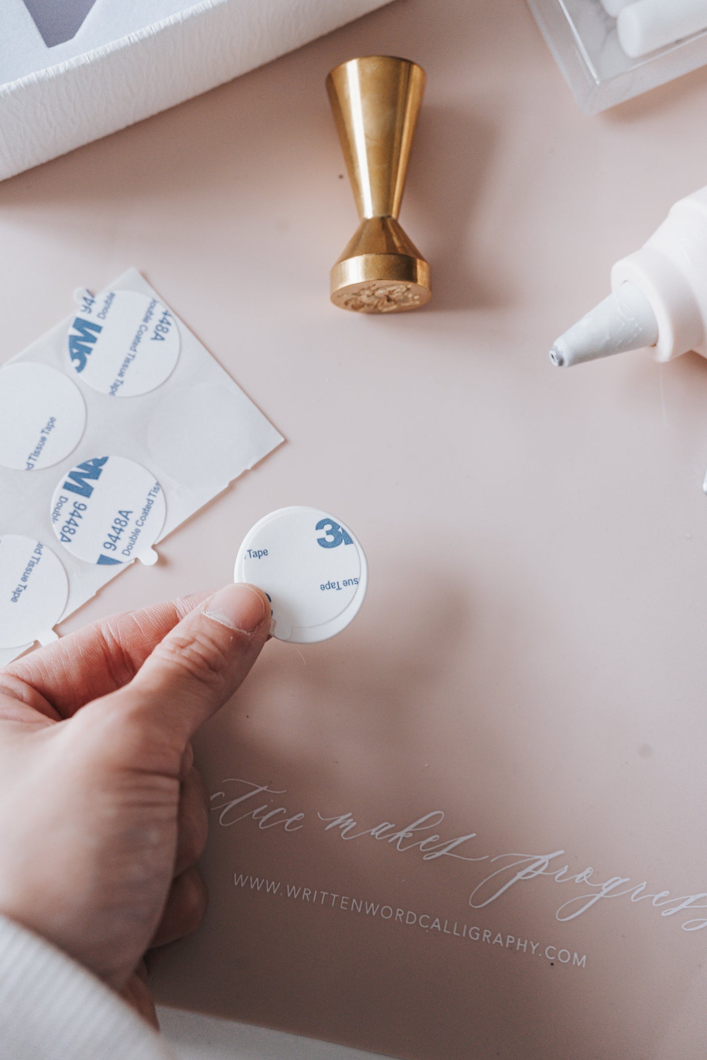 A hand holding a round adhesive seal next to a brass wax seal stamp, glue gun, and wax seal supplies on a soft pink mat.