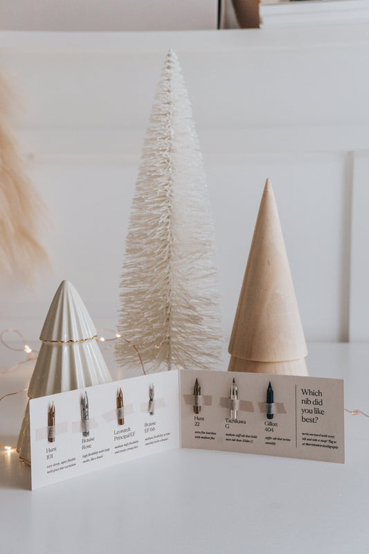 A selection of 7 calligraphy nibs displayed in a booklet next to white holiday trees