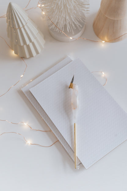 Resin calligraphy pen on slanted grid notepad surrounded by decorative trees and fairy lights