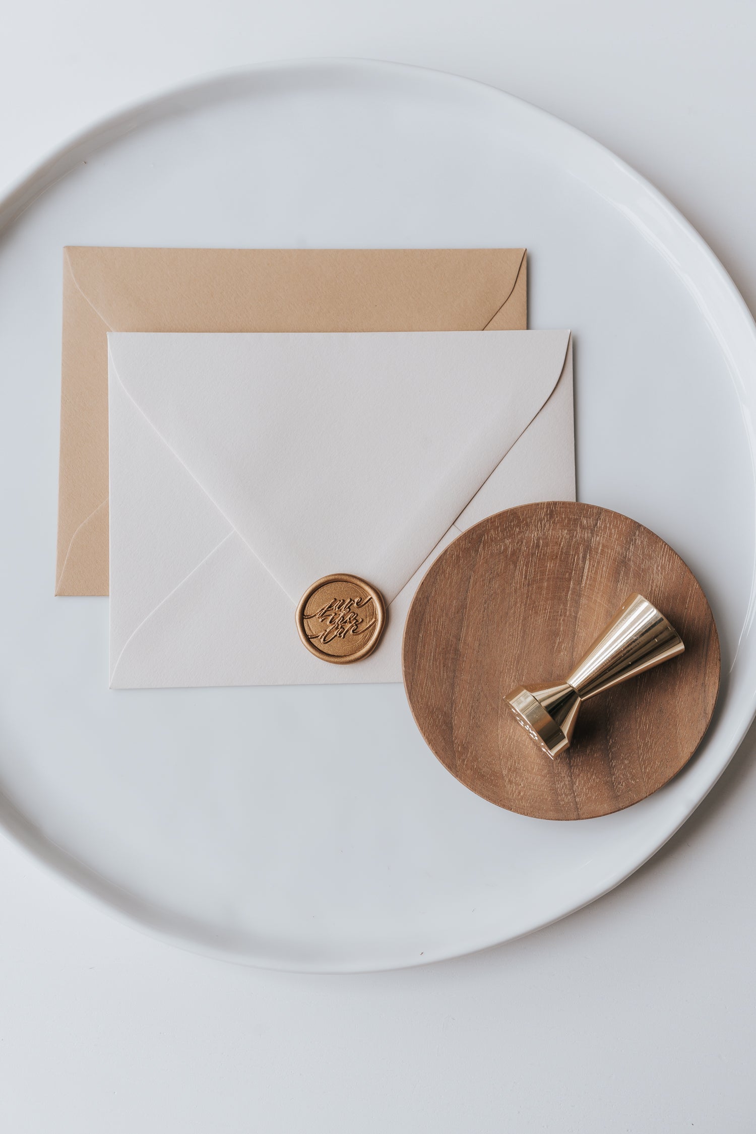 Brass wax seal stamp on a wooden dish beside envelopes with a gold save the date wax seal, styled on a white ceramic plate.