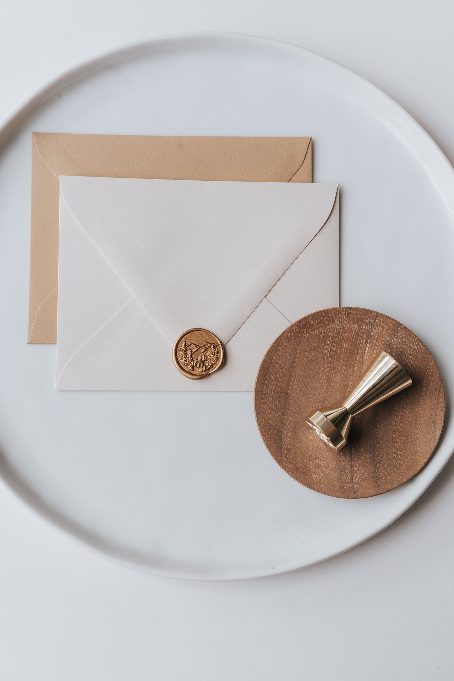 Brass wax seal stamp on a wooden dish beside envelopes with a gold trees wax seal, styled on a white ceramic plate.