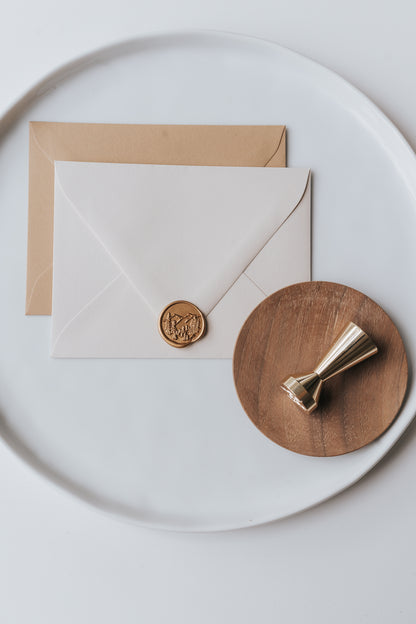 Brass wax seal stamp on a wooden dish beside envelopes with a gold trees wax seal, styled on a white ceramic plate.