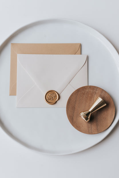 Brass wax seal stamp on a wooden dish beside envelopes with a gold birds wax seal, styled on a white ceramic plate.