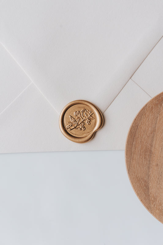 A golden birds wax seal, styled on a beige sealed envelope and a wooden dish on the side