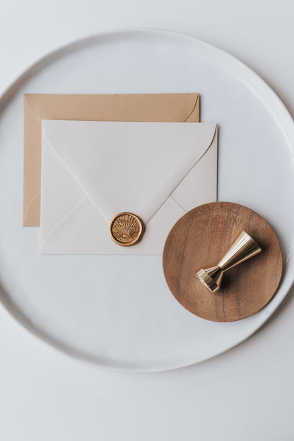 Brass wax seal stamp on a wooden dish beside envelopes with a gold sunrise wax seal, styled on a white ceramic plate.