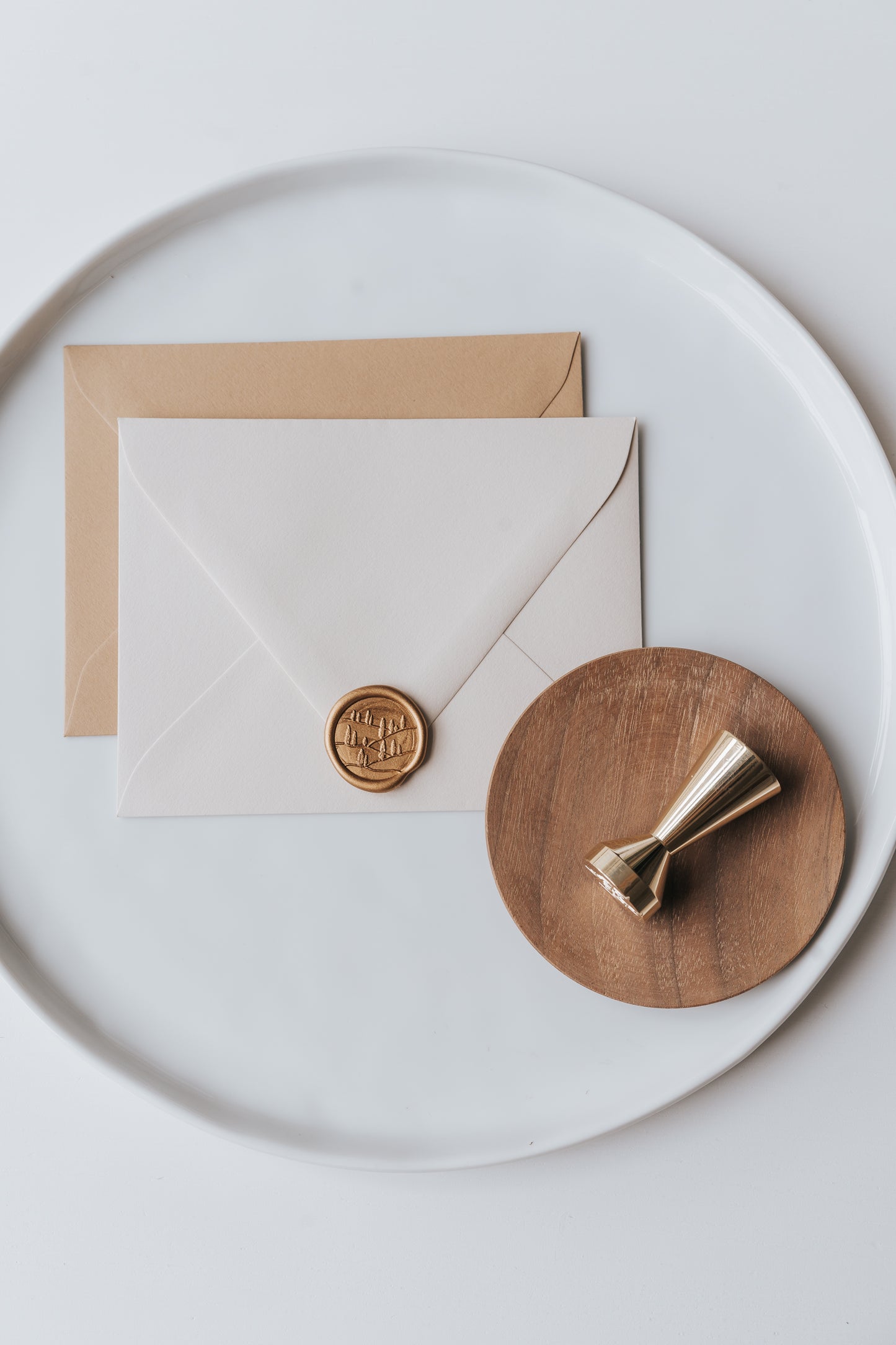 Brass wax seal stamp on a wooden dish beside envelopes with a gold Tuscany wax seal, styled on a white ceramic plate.