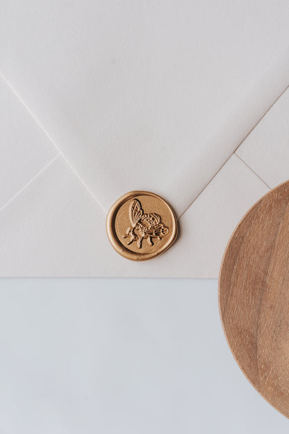 A golden honeybee wax seal, styled on a beige sealed envelope and a wooden dish on the side
