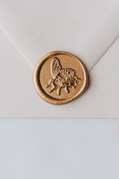 A close-up of a golden honeybee wax seal, styled on a beige sealed envelope on a white surface