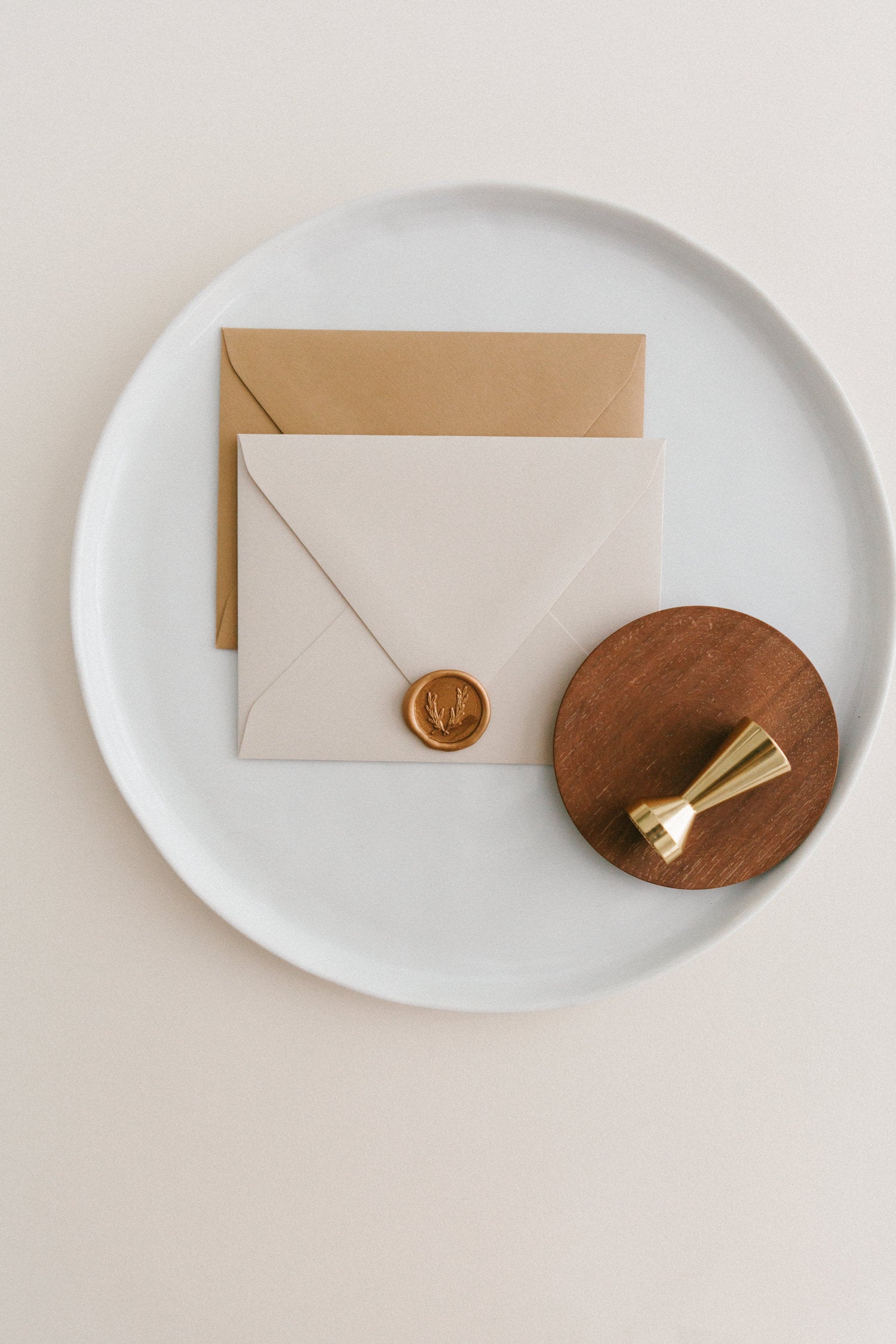 Brass wax seal stamp on a wooden dish beside envelopes with a gold olive crest wax seal, styled on a white ceramic plate.