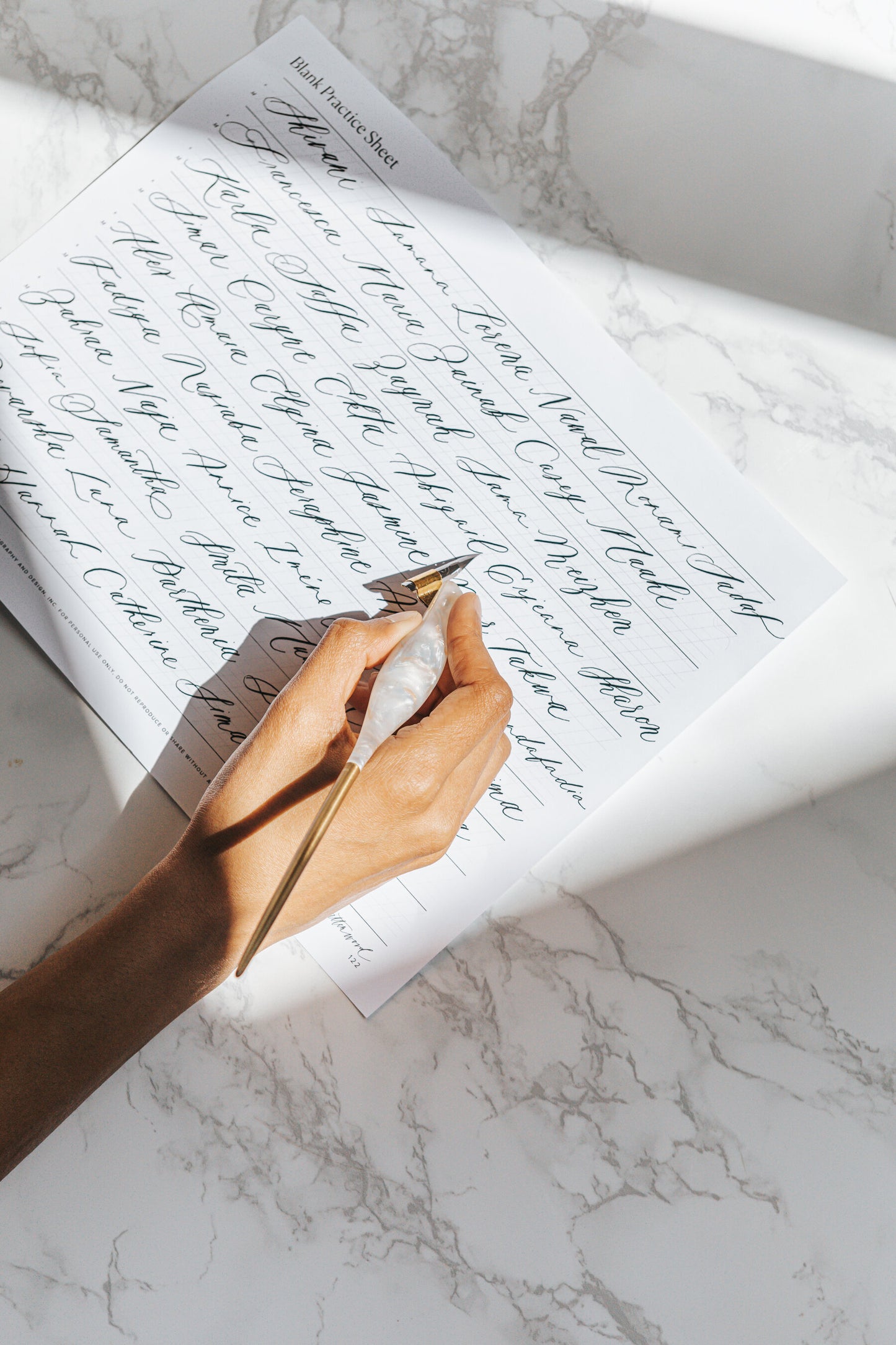A hand holding a pearl-toned oblique calligraphy pen, writing elegant cursive on a blank practice sheet on a marble surface