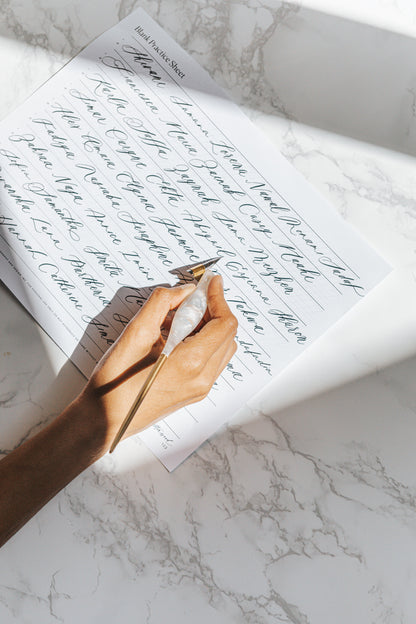A hand holding a pearl-toned oblique calligraphy pen, writing elegant cursive on a blank practice sheet on a marble surface
