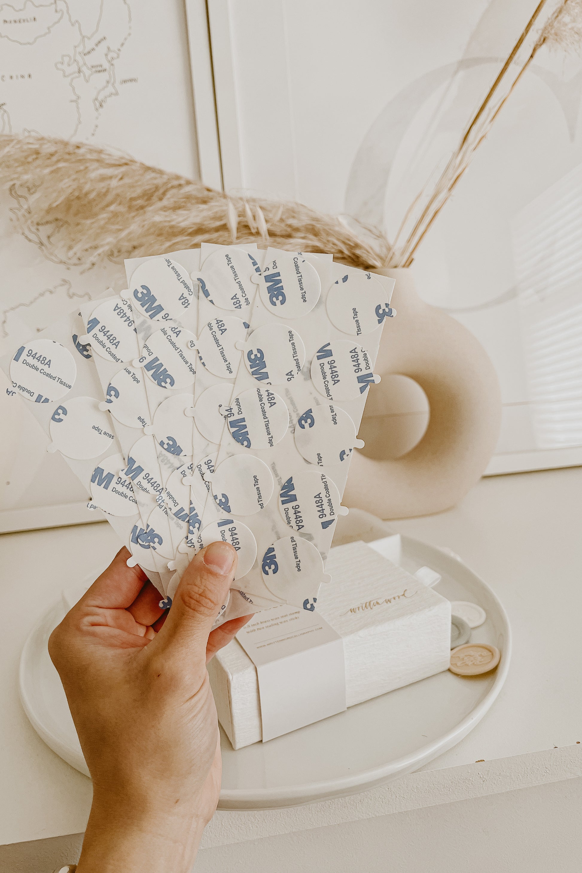Hand holding sheets of 3M double-sided wax seal adhesives against a neutral background with a vase, dried grass, and packaging.