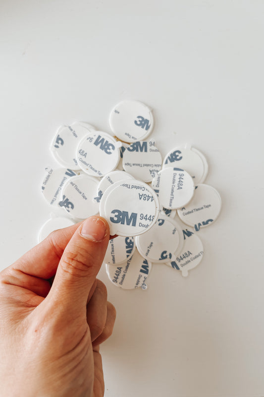 Hand holding a round wax seal adhesive with a pile of additional adhesives in the background on a white surface.