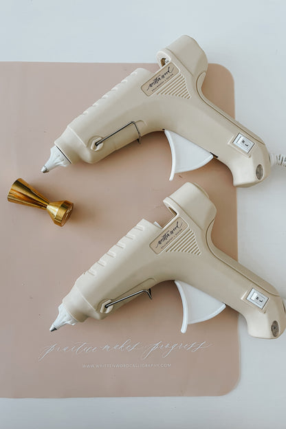 Two beige Dual-Heat Glue Guns and a brass wax seal stamp on a pink Written Word branded silicon mat