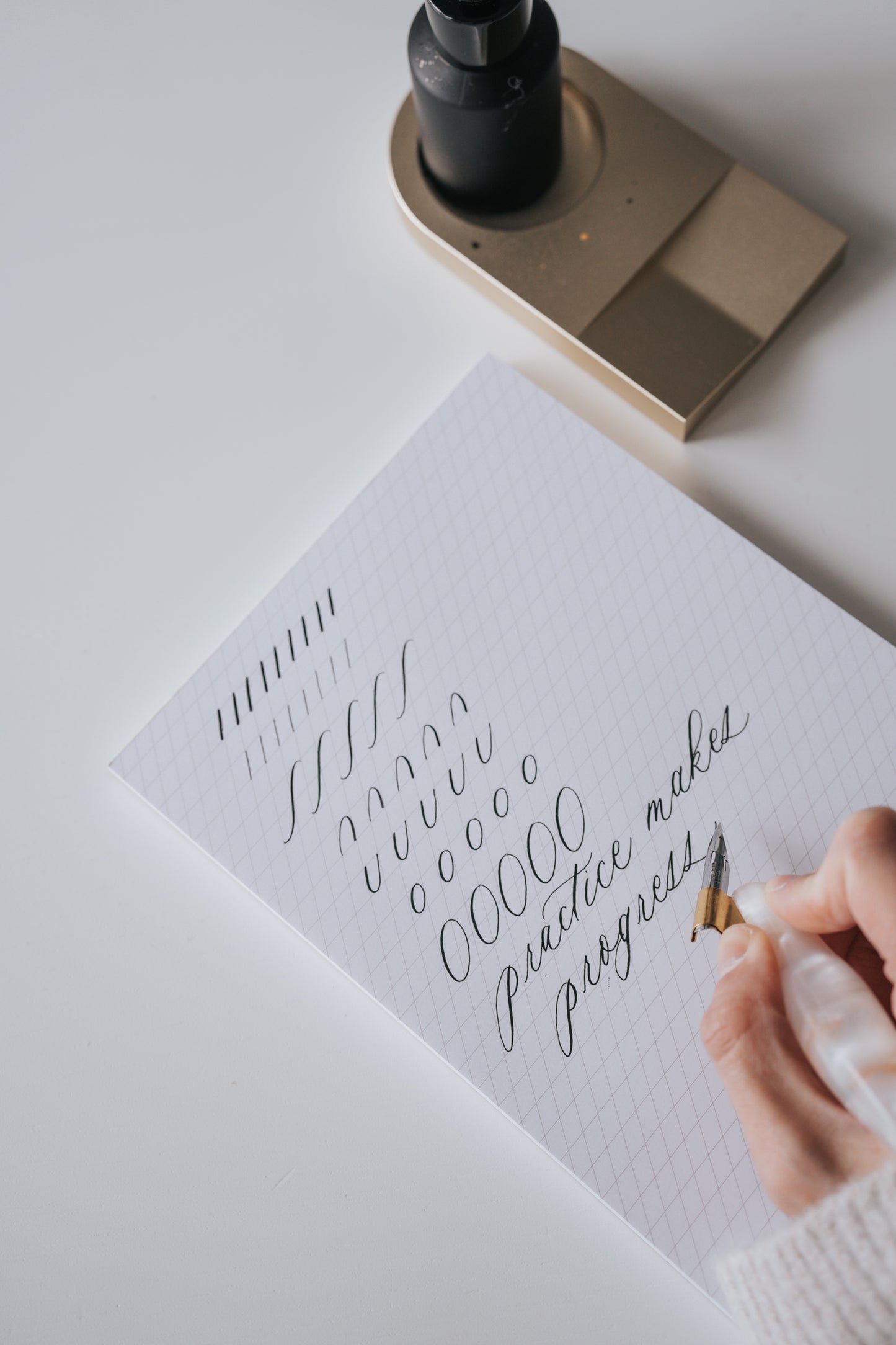 Hand practicing calligraphy on a slanted guide notepad, writing 'practice makes progress' with a pointed pen