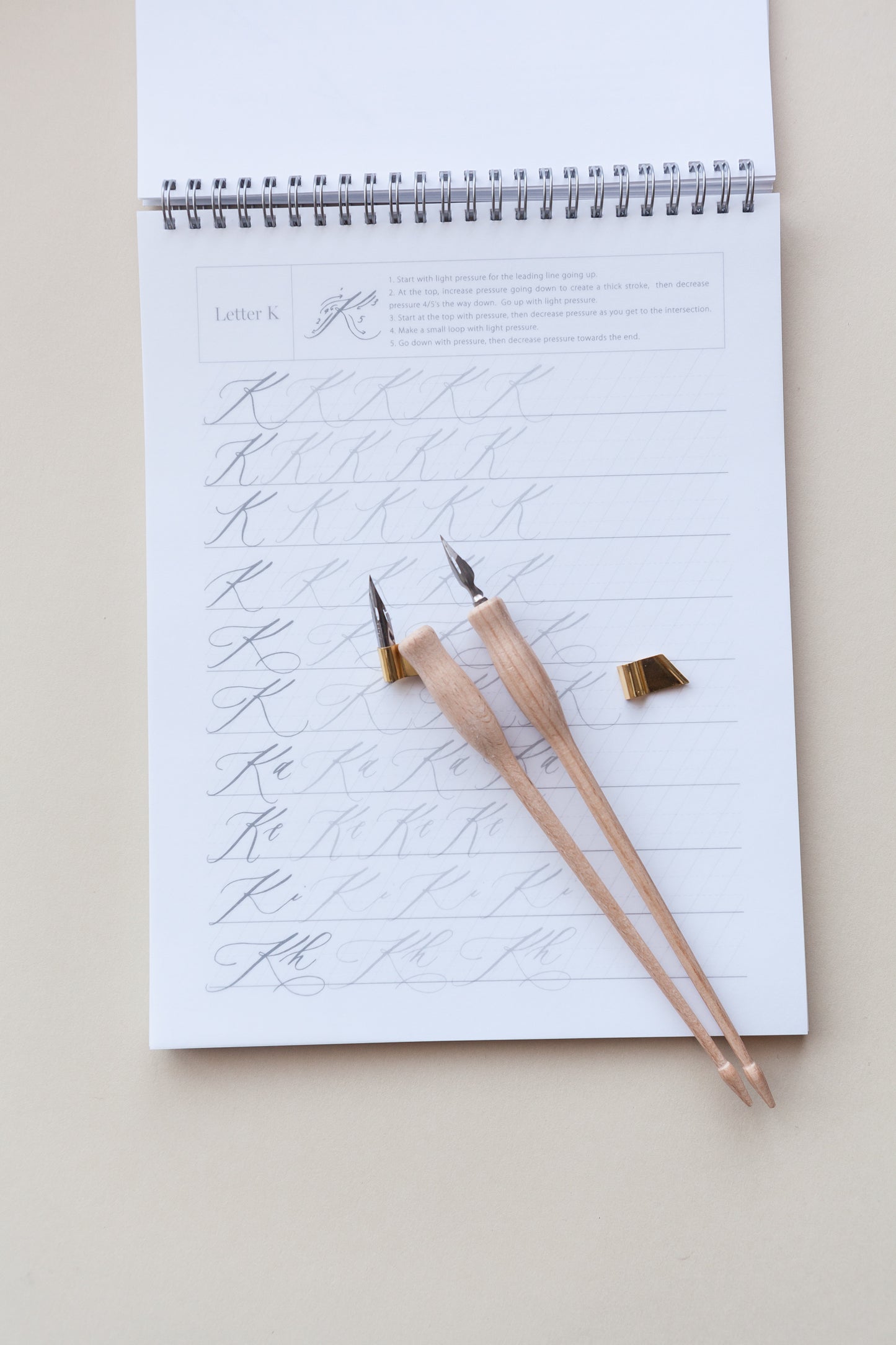 A calligraphy practice sheet featuring the letter 'K,' two wooden calligraphy pens, and a brass flange, arranged neatly