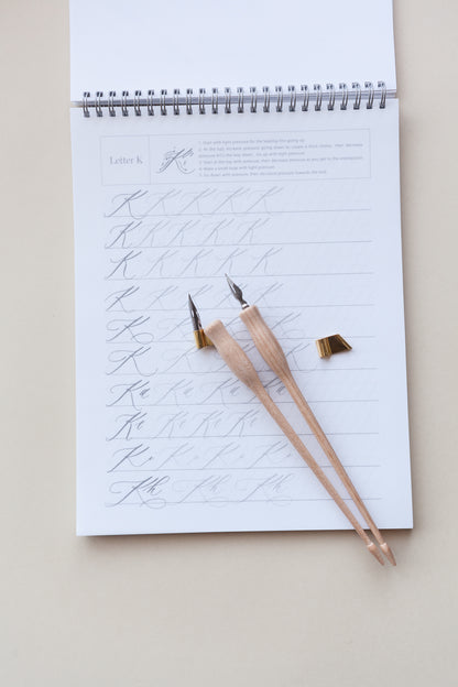 A calligraphy practice sheet featuring the letter 'K,' two wooden calligraphy pens, and a brass flange, arranged neatly