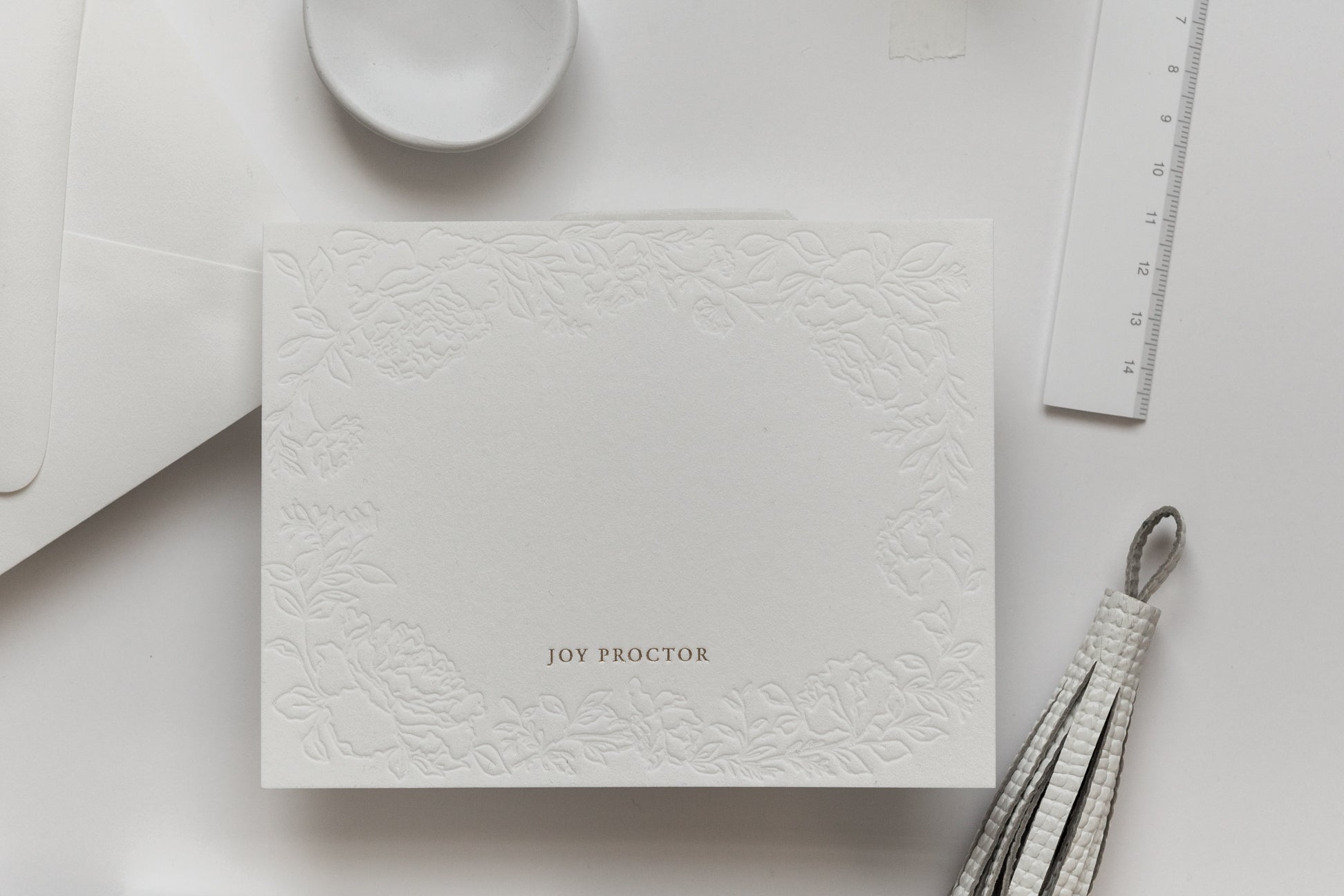 Flat lay of embossed floral card with gold foil name and neutral desk styling