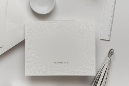 Flat lay of embossed floral card with gold foil name and neutral desk styling