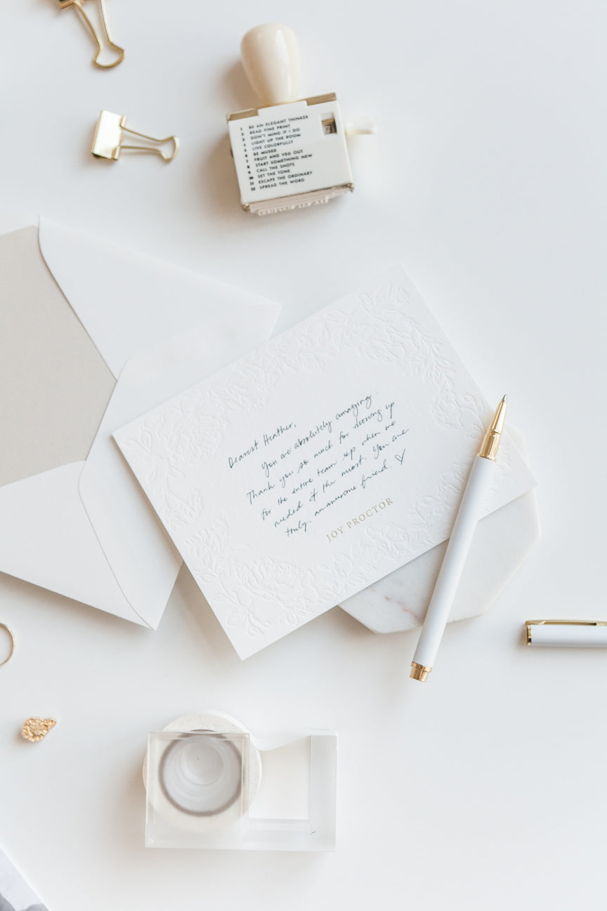 Neutral flat lay with envelope, embossed card, pen, and gold desk accessories